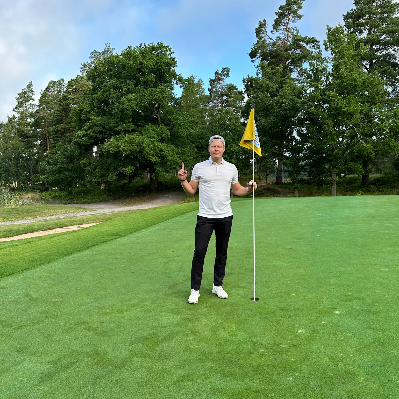 Man i vit pikétröja och svarta byxor står på en golfgreen, håller i flaggstången och visar ett tecken med handen. Skog i bakgrunden.