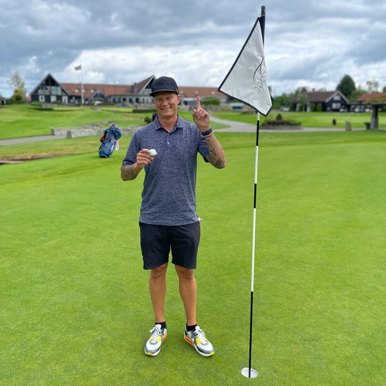 Man holder en golfbold og peger opad ved siden af et hul med flag på en golfbane, klubhus ses i baggrunden.