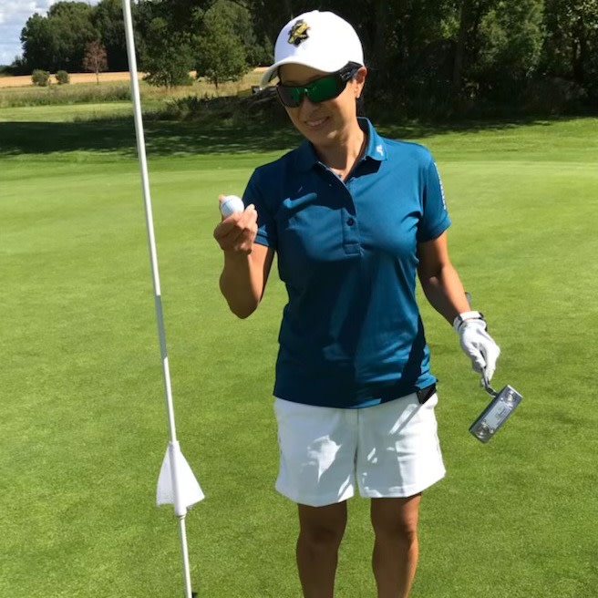 Kvinde i golfbeklædning holder en golfbold og en putter ved siden af et hul på en grøn golfbane i solskin.