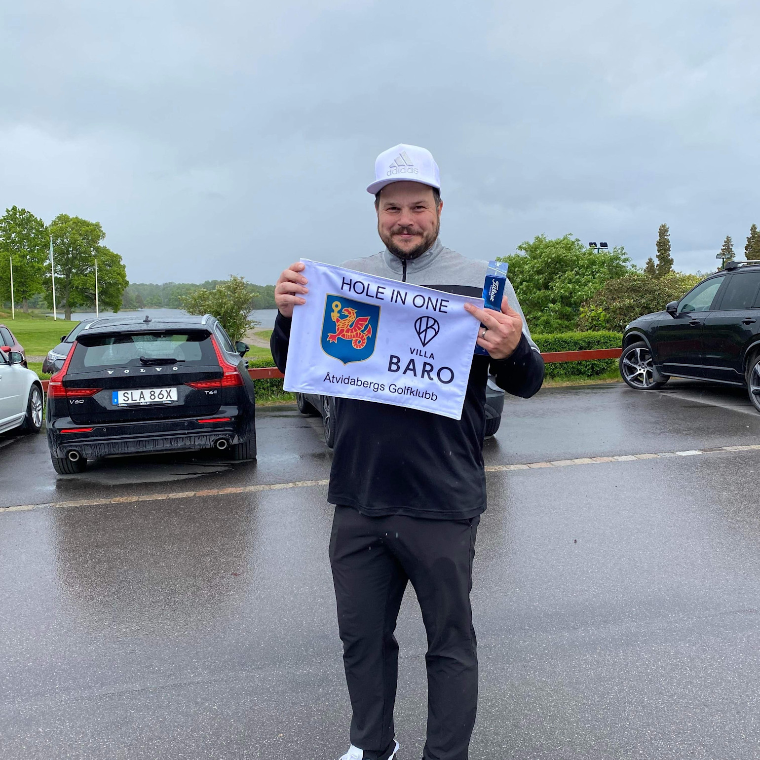 Man holder et flag med teksten "HOLE IN ONE", "VILLA BARO", "Åtvidabergs Golfklubb" på en parkeringsplads, omgivet af biler og grønne omgivelser.
