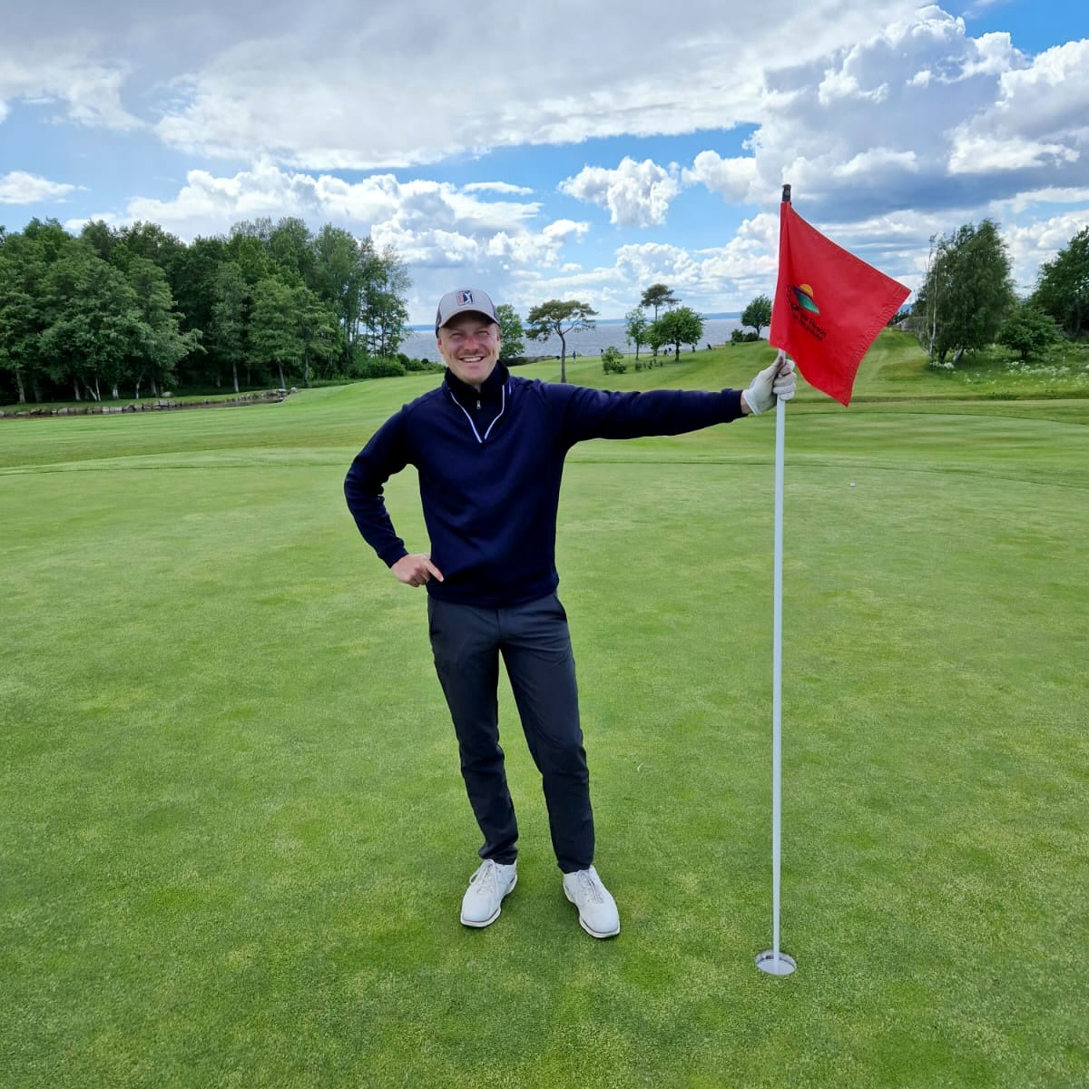 Man smiler og holder en golfstang med et rødt flag på en golfbane, omgivet af grønt græs og træer under en overskyet himmel.