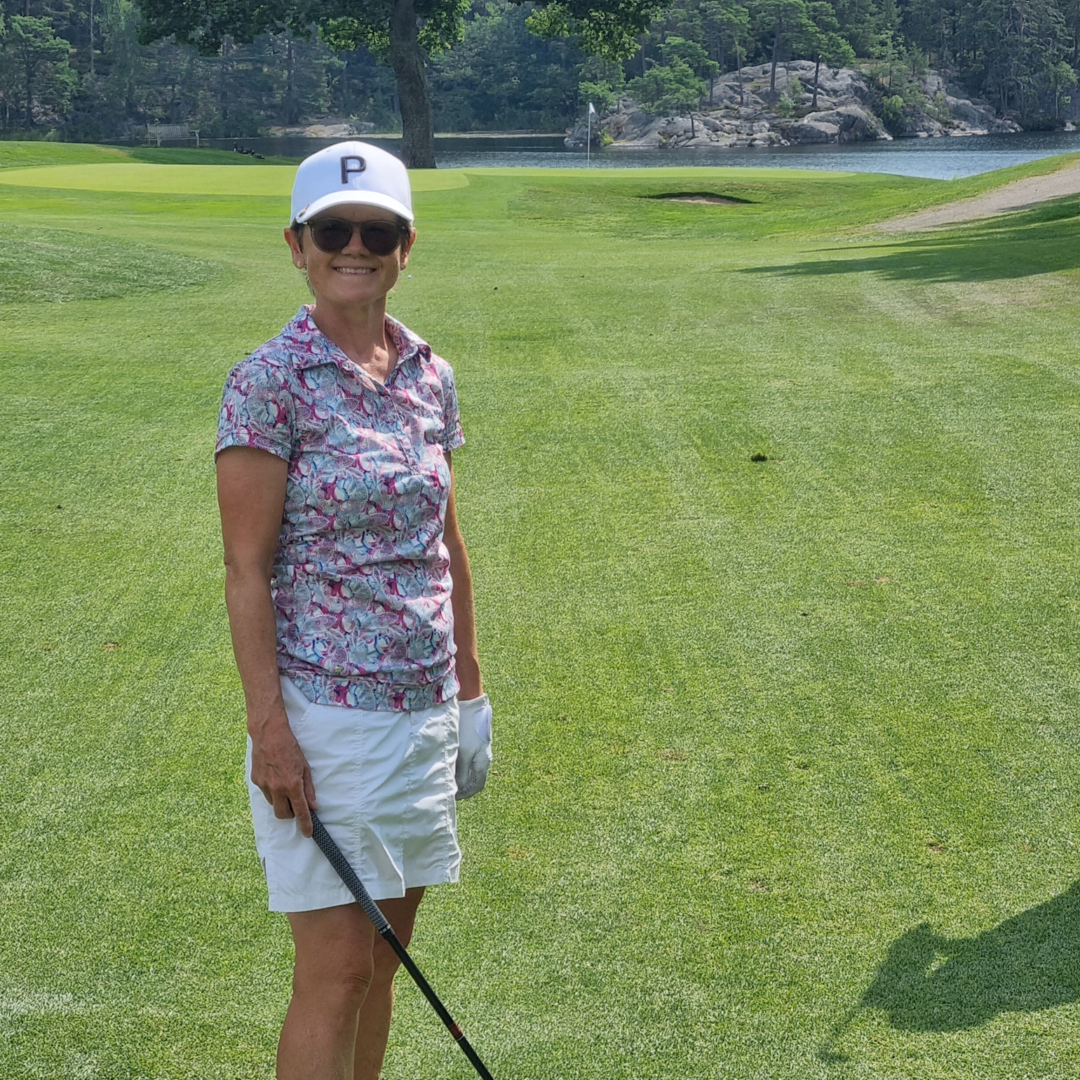 Kvinde med golfkølle står på en grøn golfbane, bærer hvid kasket med bogstavet P, solbriller og blomstret trøje.