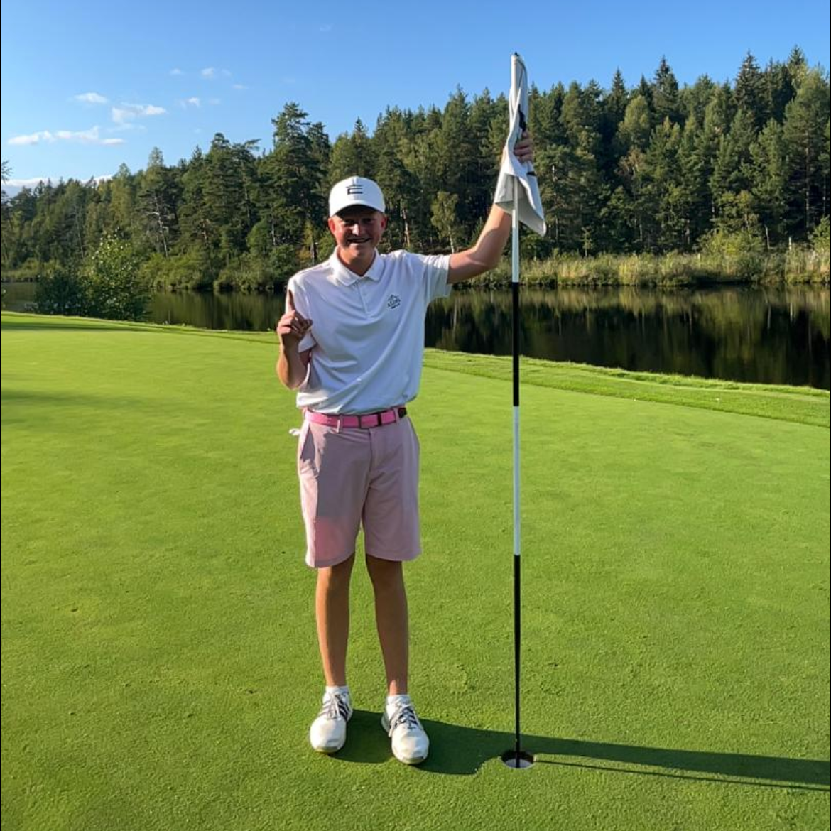 Ung person i vit pikétröja och rosa shorts står på en golfgreen, håller i flaggstången och visar ett finger, med skog och sjö i bakgrunden.