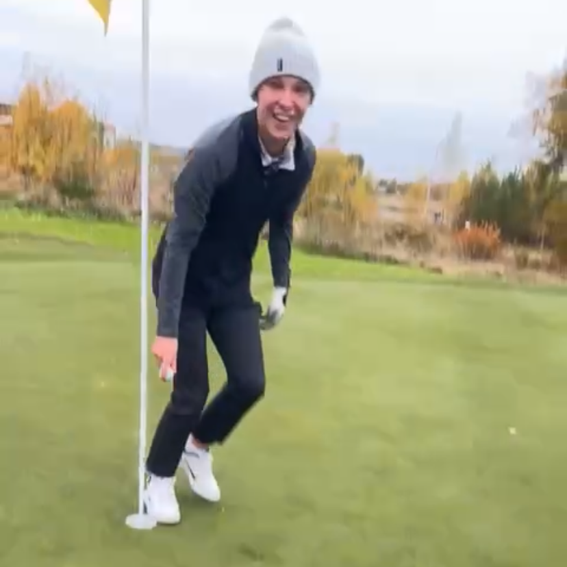 Leende person i mössa och golfkläder böjer sig vid en flaggstång på en golfgreen under höst med träd i bakgrunden.