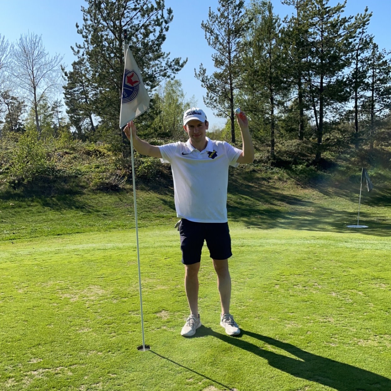 Man i vit pikétröja och keps står vid en golfhålsflagga, håller upp en golfboll och firar på en solig golfbana med träd i bakgrunden.