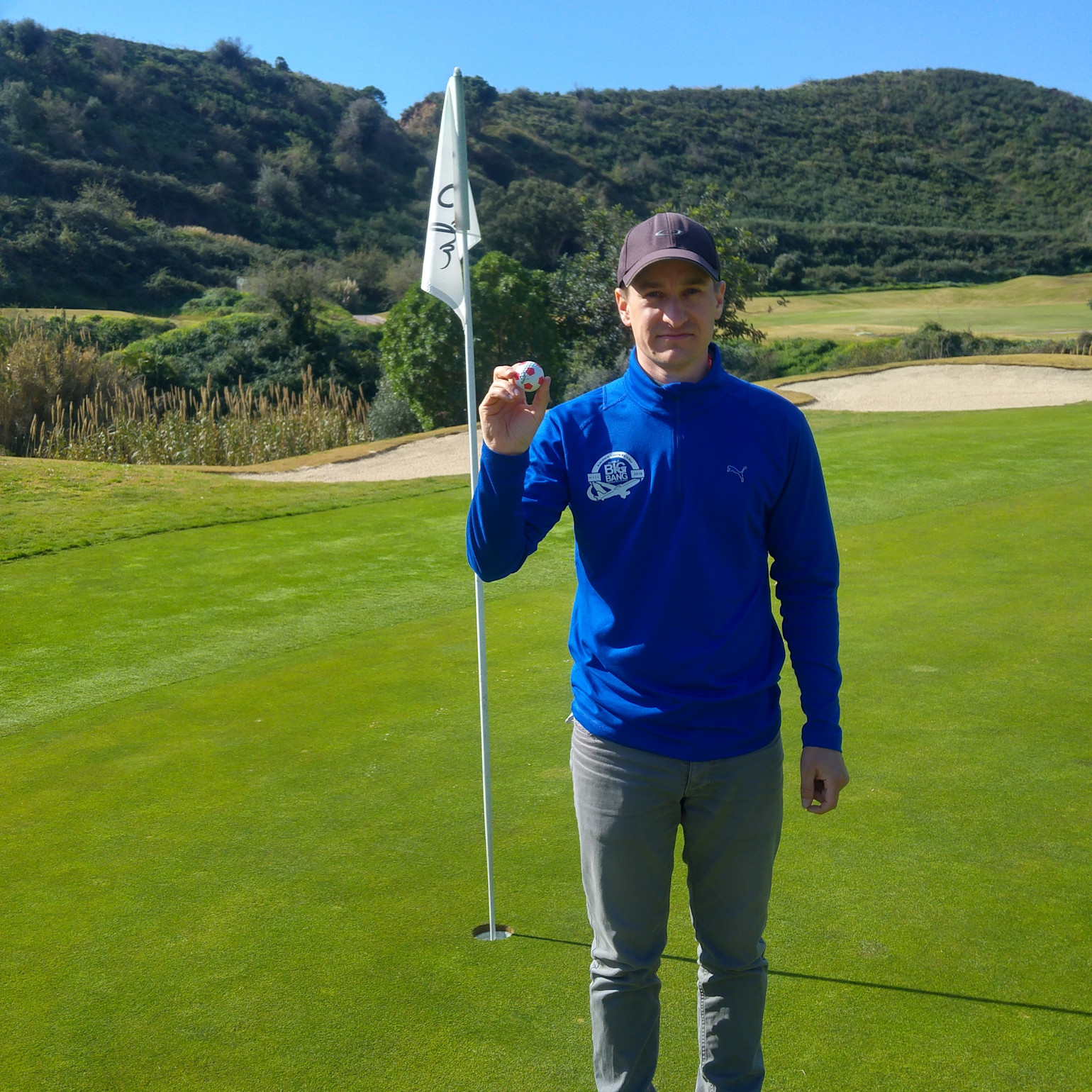 Mand på blå trøje og kasket holder en golfbold op ved siden af en flagstang på en golfbane med grønne bakker i baggrunden.
