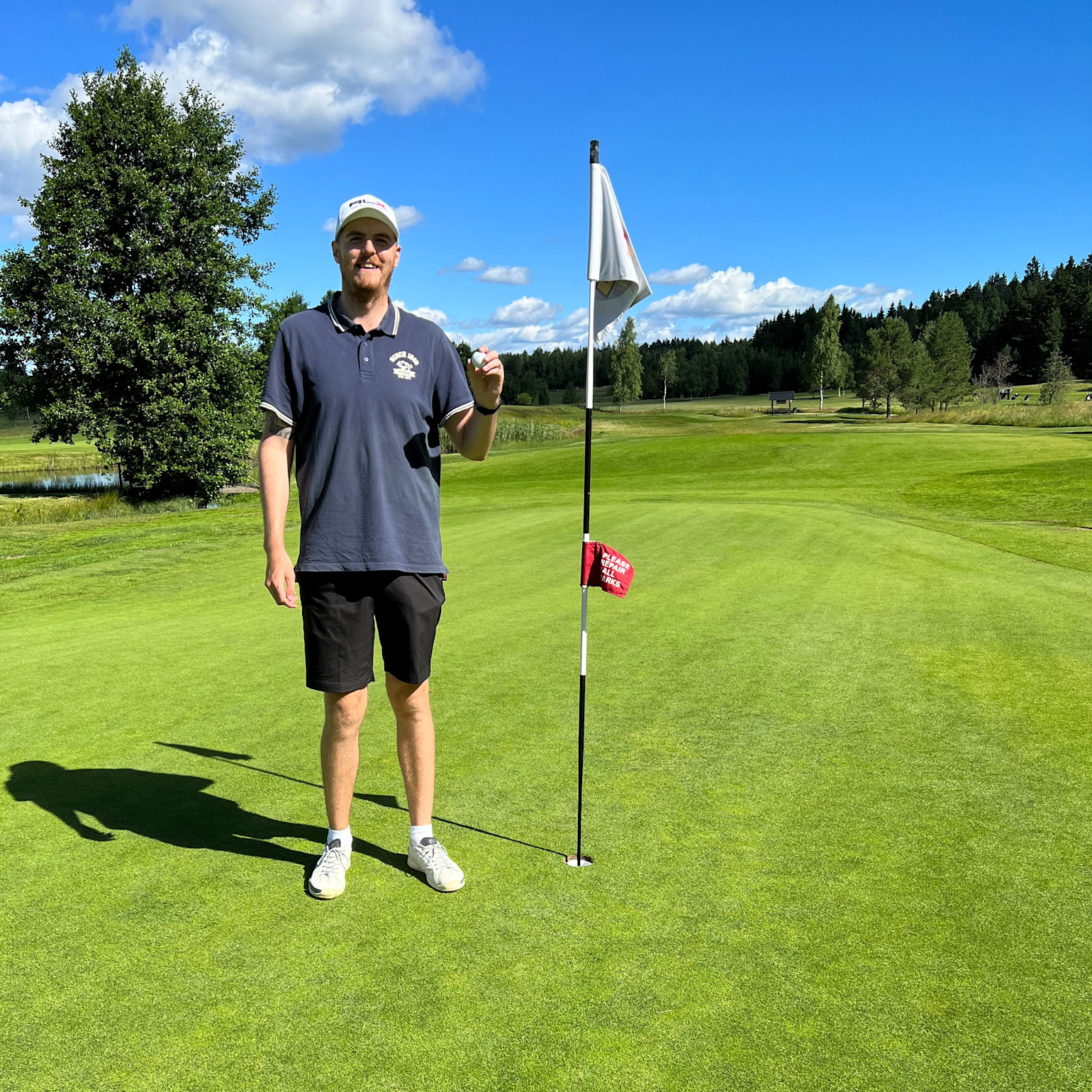 Mand på mørkeblå poloshirt og shorts står ved siden af en flagstang på en golfgreen, holder en golfbold op, solrig dag med blå himmel.
