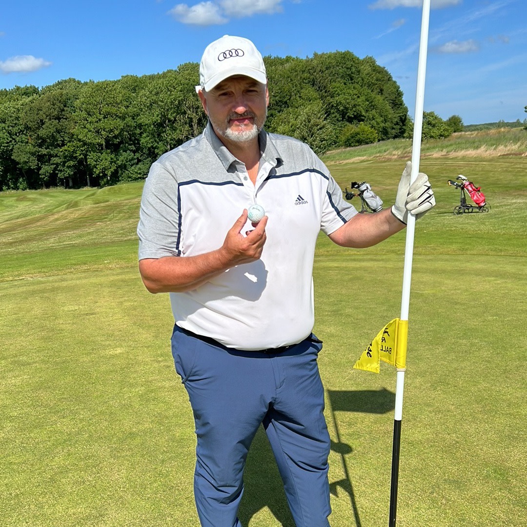 En mand i golfbeklædning holder en golfbold og en flagstang på en golfbane med grønt græs og træer i baggrunden.