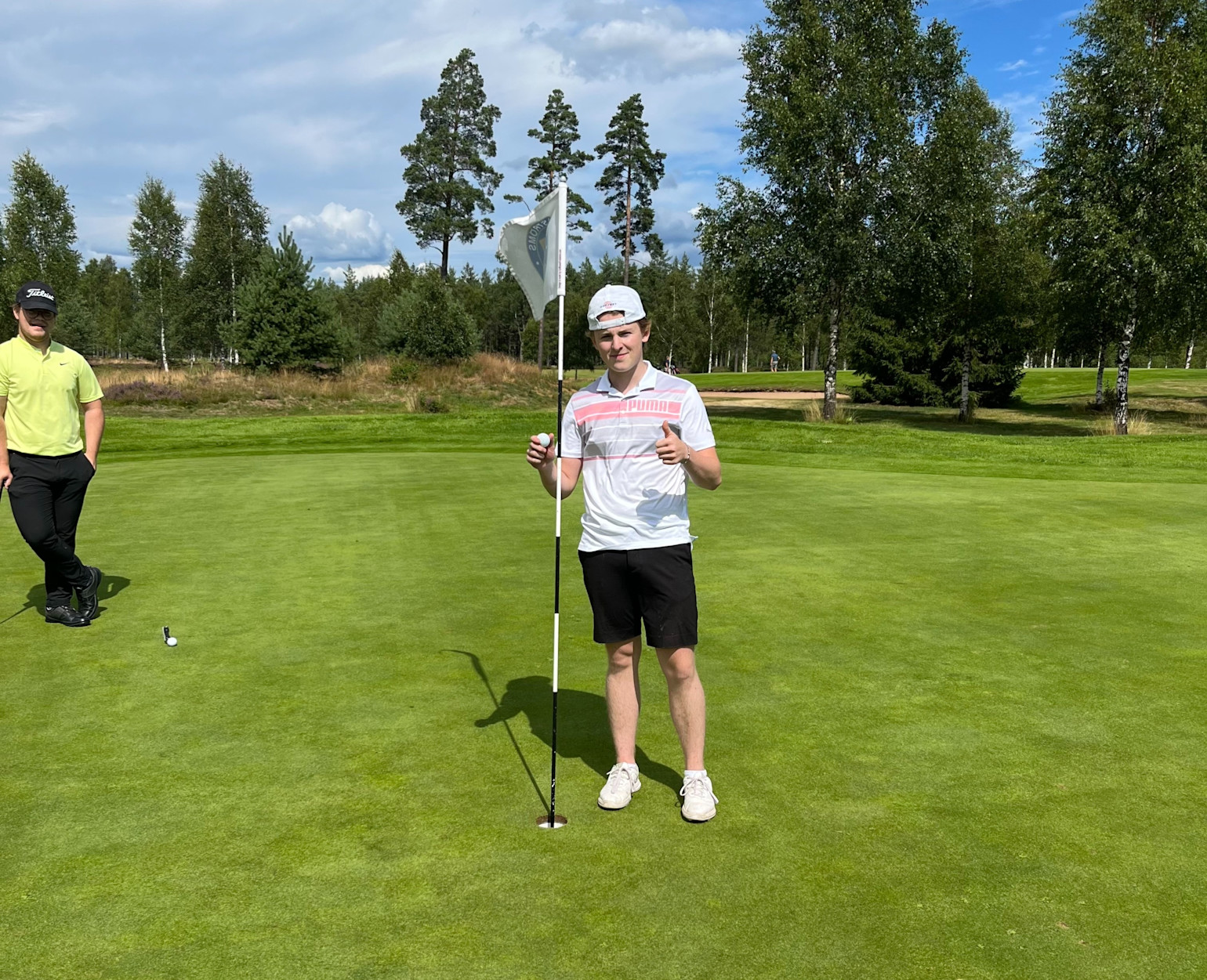 Ung mand i hvid kasket og Puma-trøje holder en golfbold og flagstang på greenen, giver tommelfingeren op, træer i baggrunden.