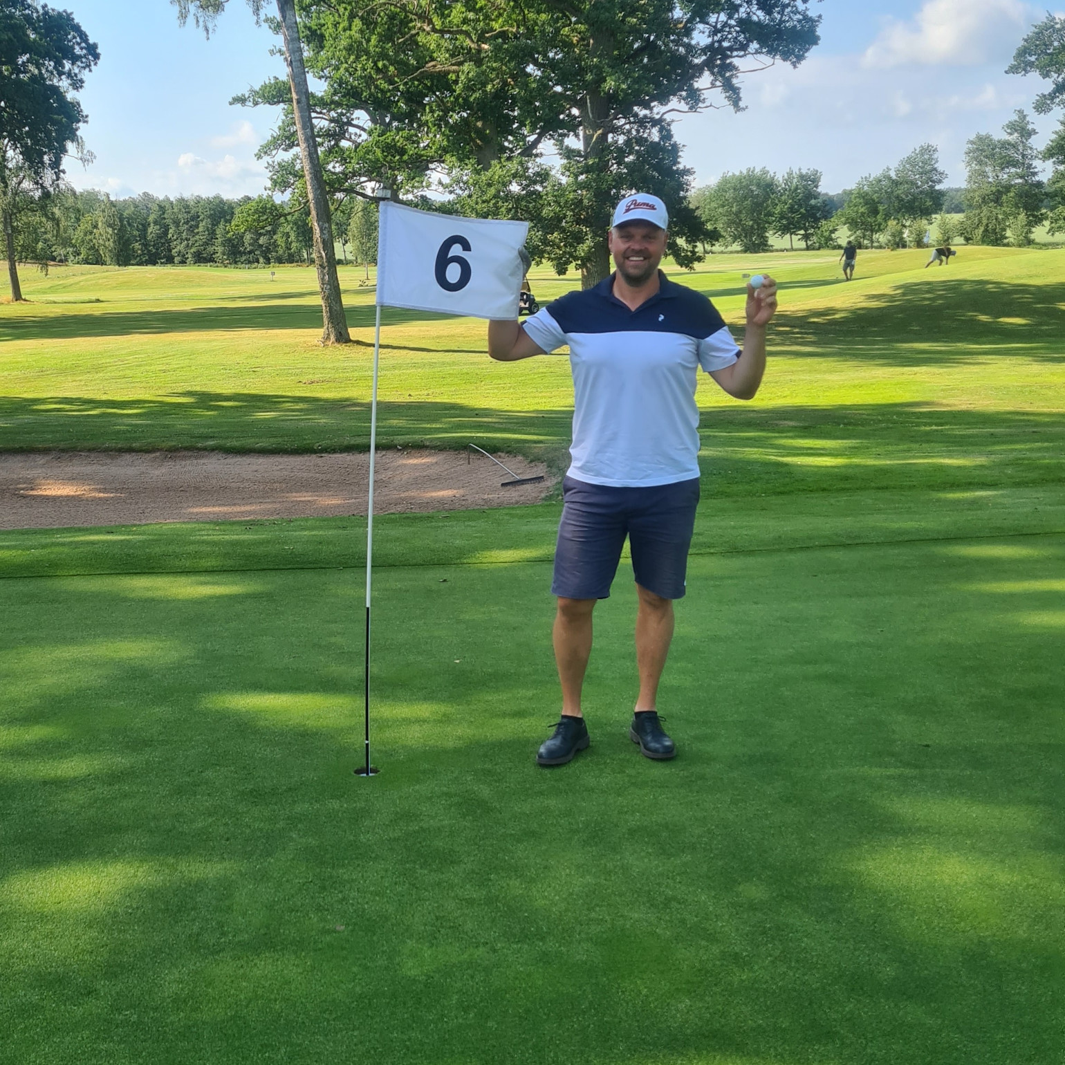 Man holder en golfbold og står ved siden af et flag med tallet 6 på en golfbane på en solrig dag.