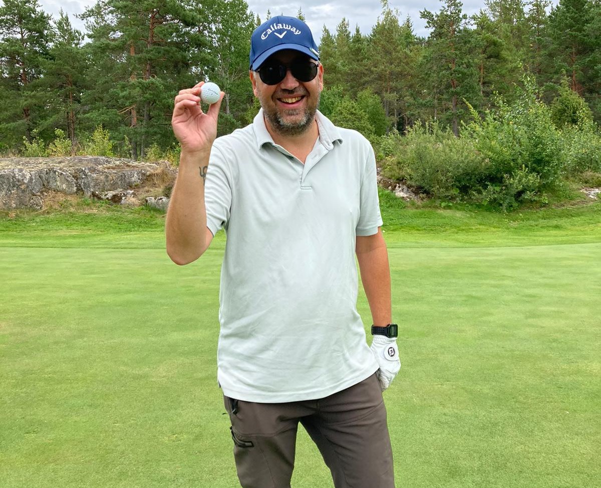 Smilende mand med solbriller og blå kasket holder en golfbold op på en golfbane omgivet af træer og græs.