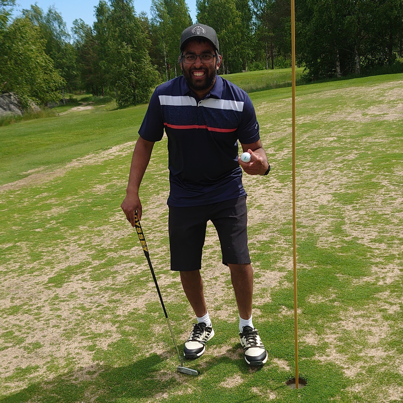 Man smiler og holder en golfbold og en putter ved siden af et hul på en golfbane omgivet af grønne omgivelser.
