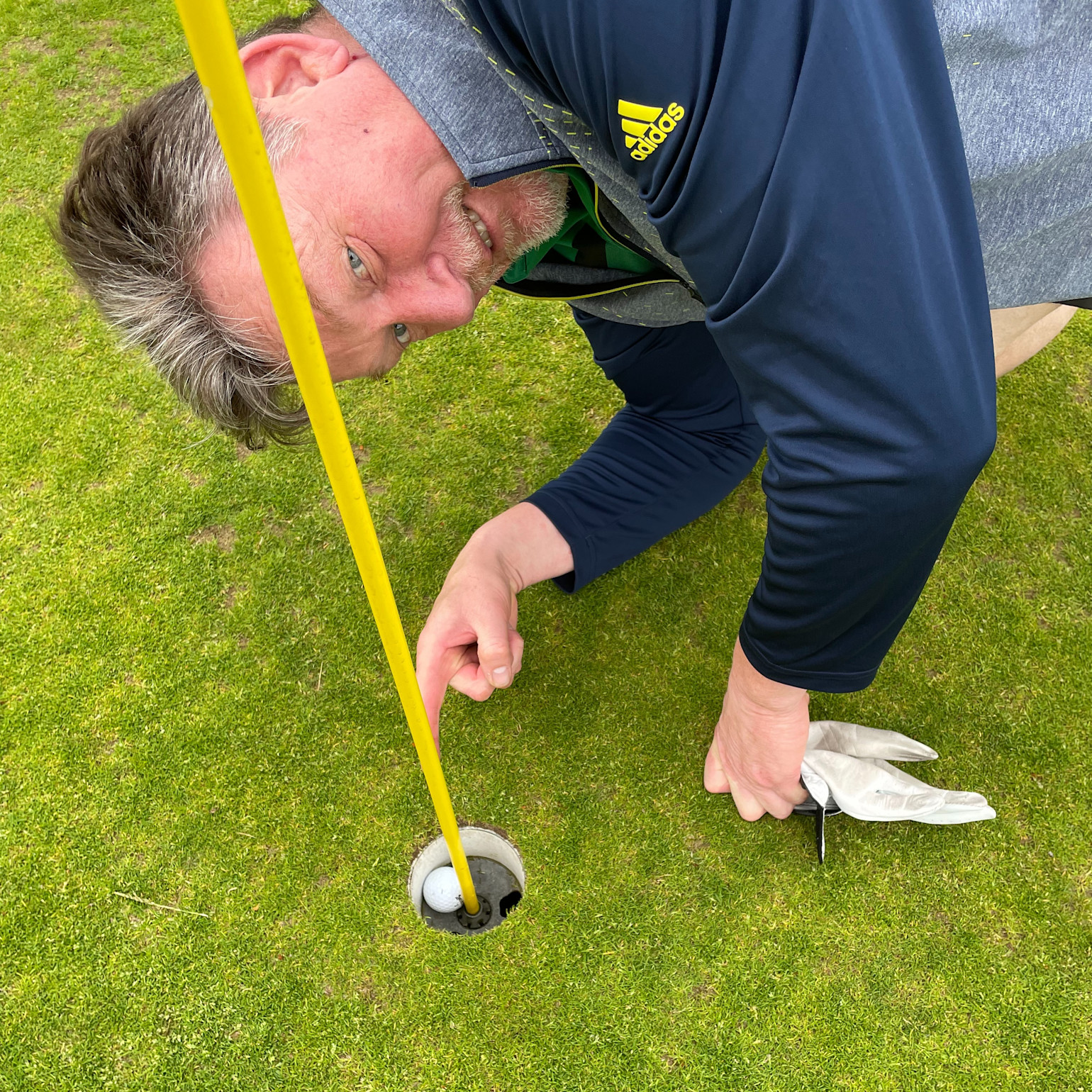 Man bøjer sig ned over et golfhul, peger på en golfbold i hullet ved siden af en gul flagstang på en grøn golfbane.