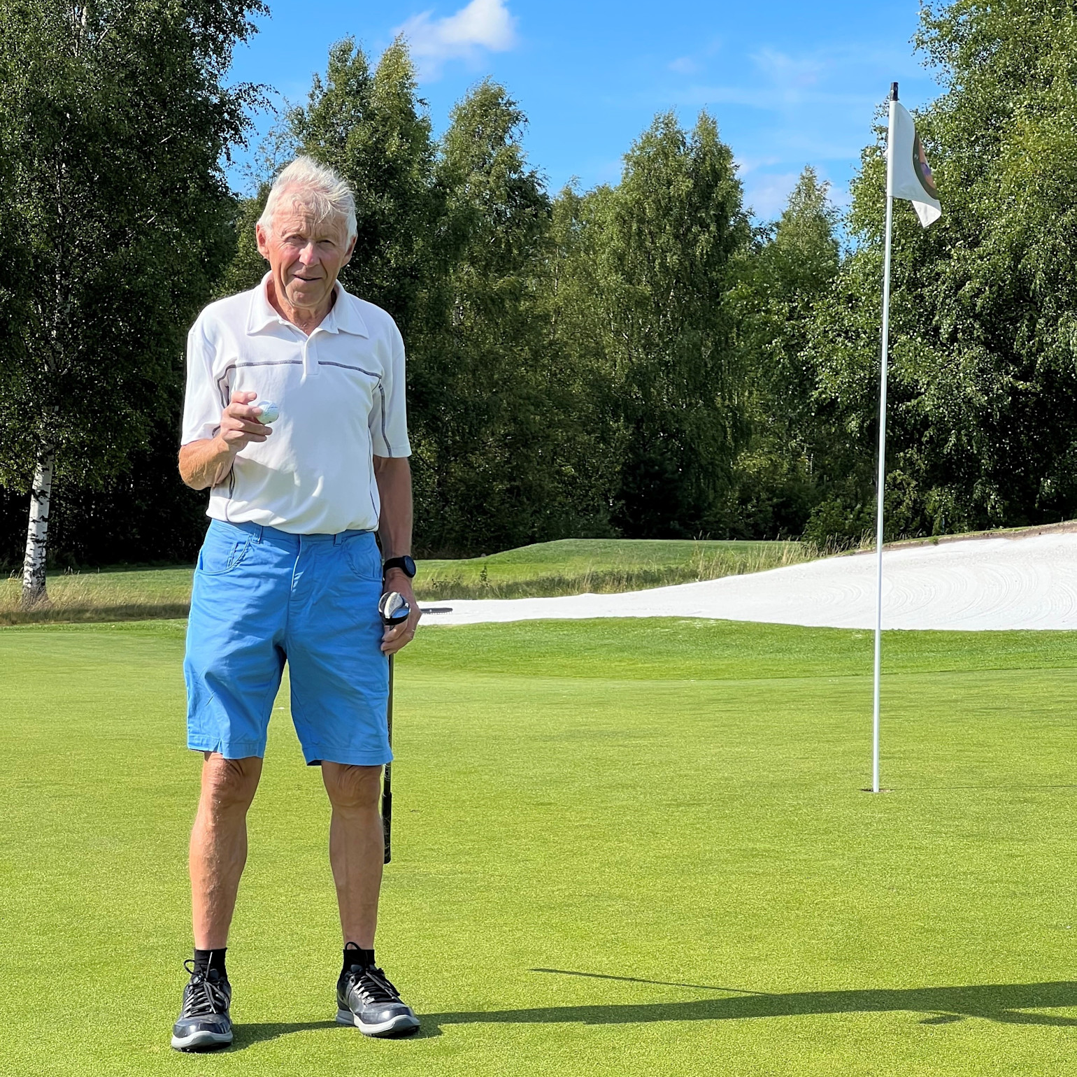 Ældre mand i hvid poloshirt og blå shorts holder en golfbold og en kølle på en green tæt ved flagstangen på en solrig dag med træer i baggrunden.