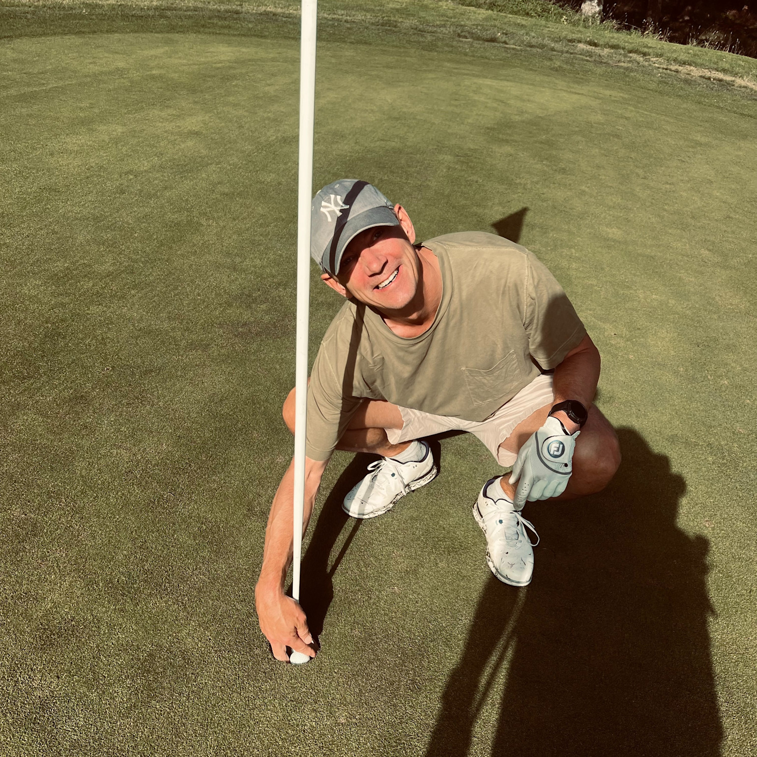 Man i keps hukar sig vid golfhål, håller i flaggstången och ler på en golfgreen i solsken.