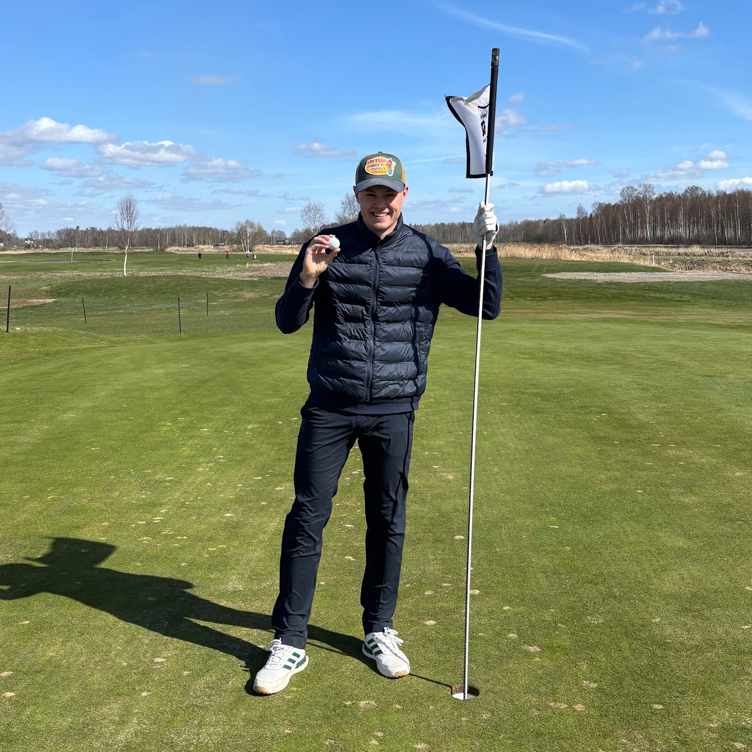 On tient une balle de golf et un drapeau sur un green de golf par une journée ensoleillée, avec des arbres et un ciel bleu en arrière-plan.