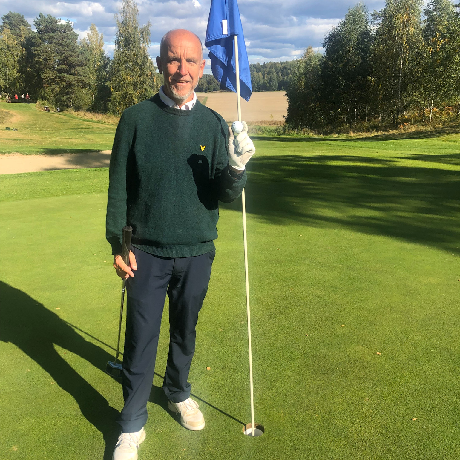 Man håller en golfboll och flaggstång vid ett hål på en golfbana, omgiven av grönska och träd i solsken.