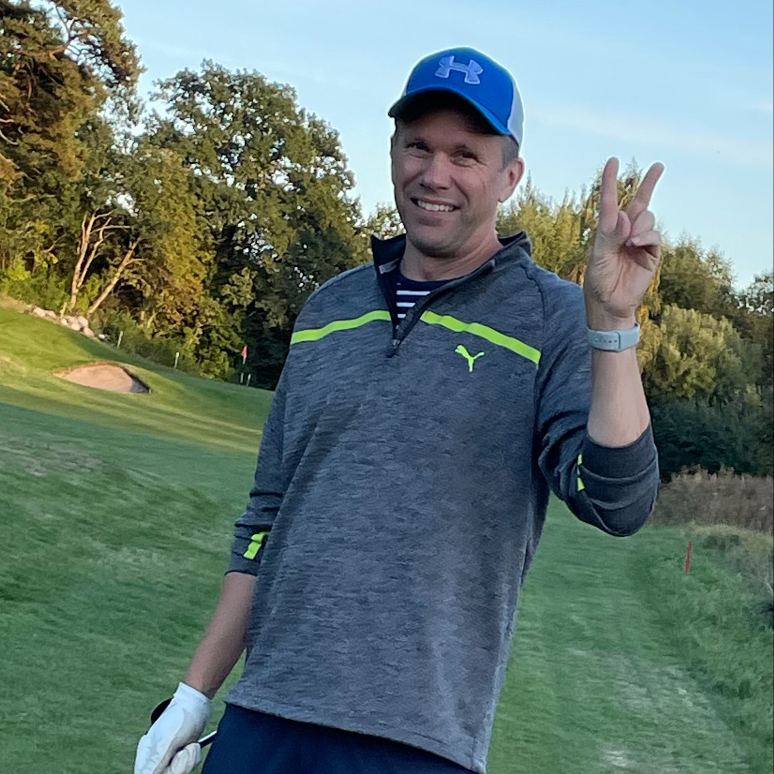 En mand iført golfbeklædning laver et sejrstegn med den ene hånd og står smilende på en golfbane omgivet af træer.