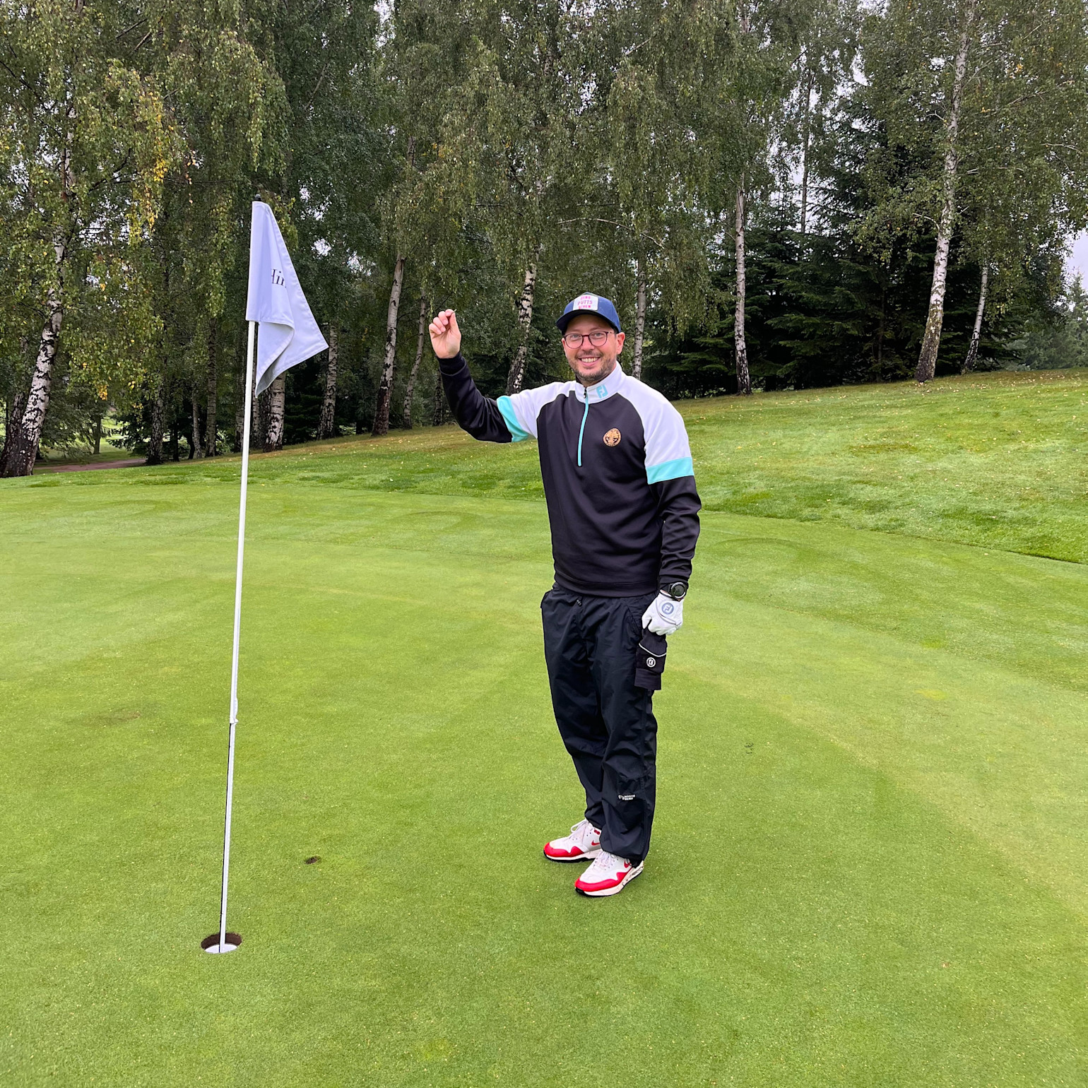 En mand iført golfbeklædning holder en golfbold op ved siden af et hul med flag på en golfbane omgivet af træer.