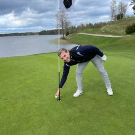 Man samler en golfbold op fra hullet på en golfbane ved en sø under en overskyet himmel.