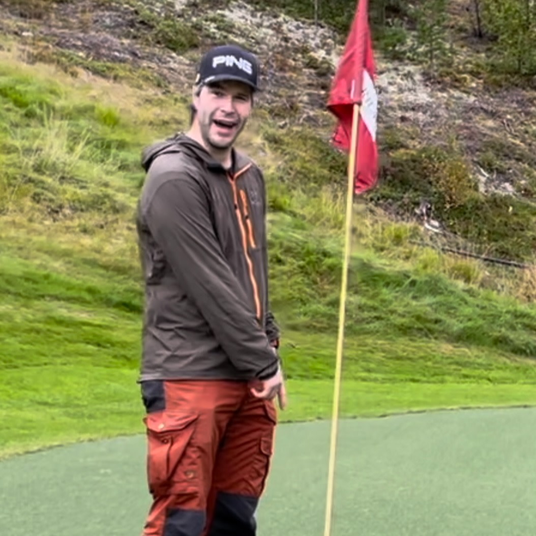 Mand med mørk jakke og rød-sorte bukser står ved siden af et golfhulflag på en golfbane, smiler og peger mod jorden.
