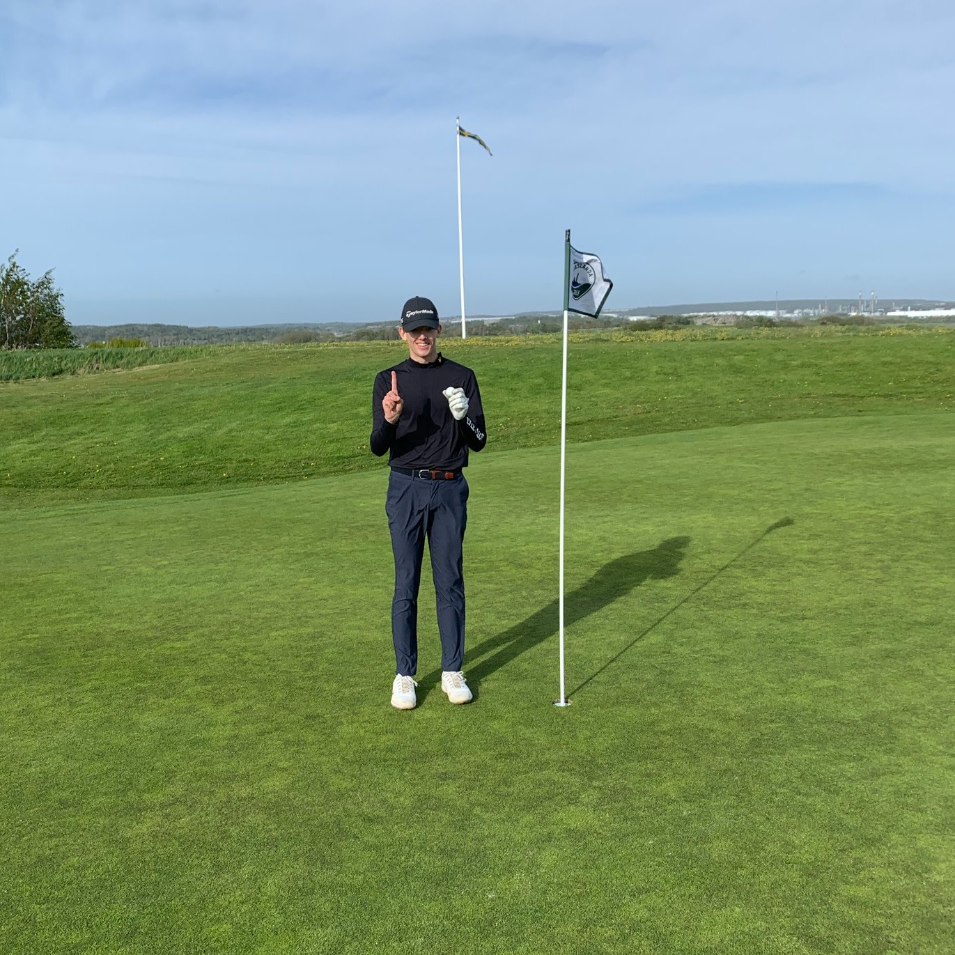 En person iført golfbeklædning står ved siden af en flagstang på en golfgreen og holder én finger op, med græs og blå himmel i baggrunden.