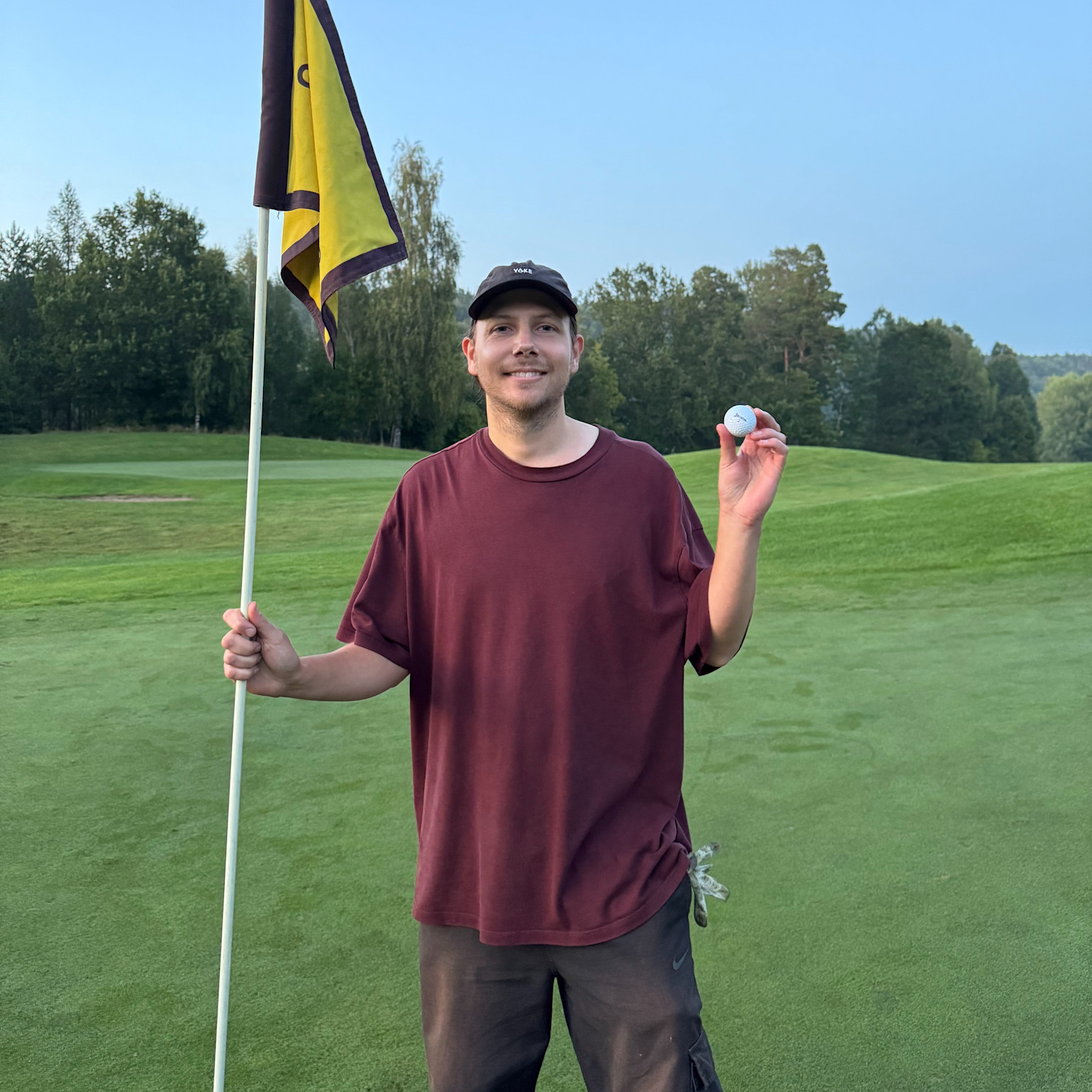 Man in a burgundy t-shirt holding a golf club flag in one hand and a golf ball in the other on a golf course with grass and trees.