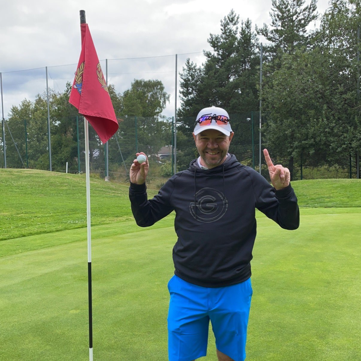 Mand i sporttøj holder en golfbold op og viser én finger ved siden af et hul med rød flag på en golfbane.
