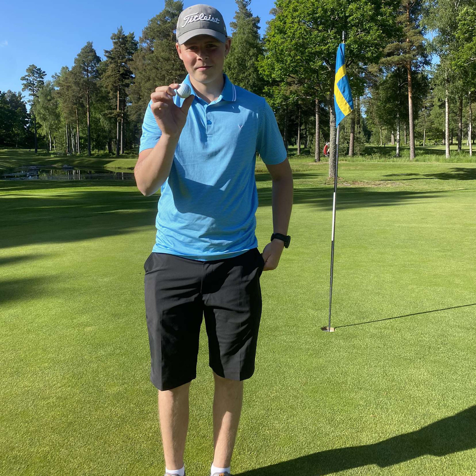 Ung mand i blå poloshirt og kasket holder en golfbold op på en green, med det svenske flag i baggrunden.