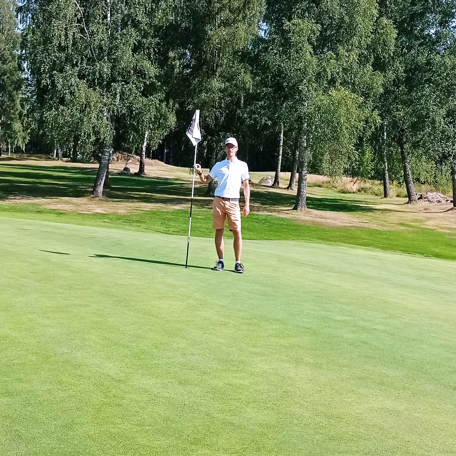 En person står på en golfgreen og holder i flagstangen, omgivet af grønt græs og træer i baggrunden.