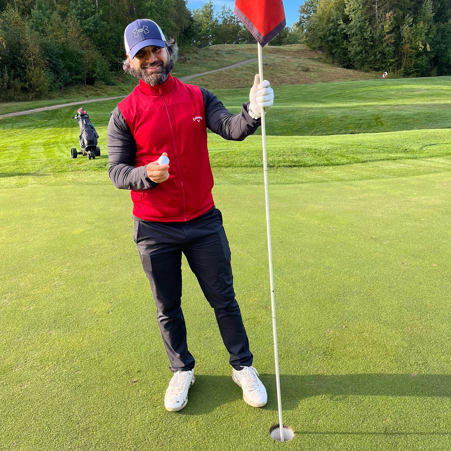 Man i röd väst och blå keps håller en golfboll och flaggstång på en golfgreen, med golfvagn och träd i bakgrunden.