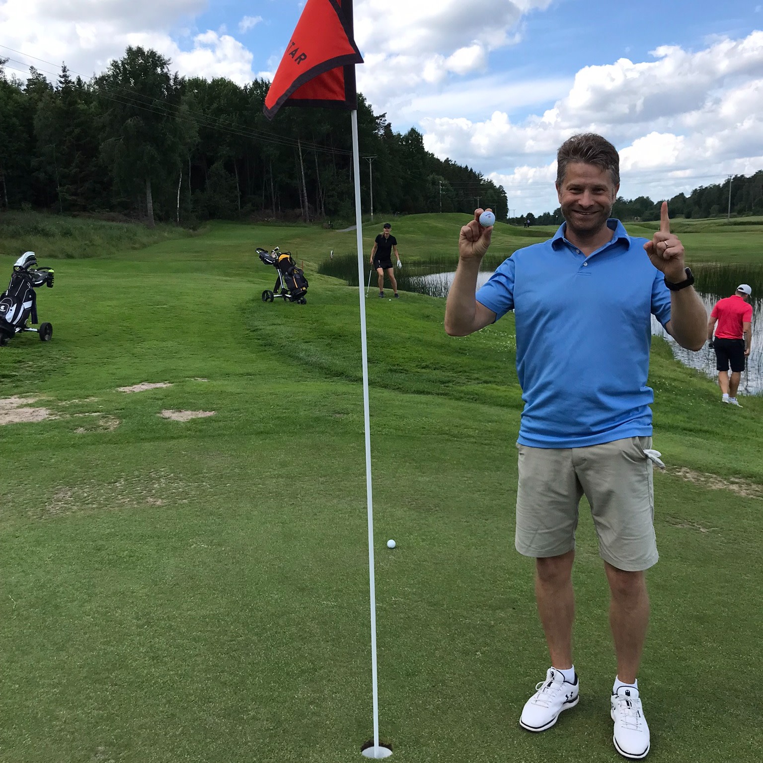 Mand iført blå poloshirt og beige shorts holder en golfbold op og peger opad ved siden af et golfhul med flag på en golfbane.