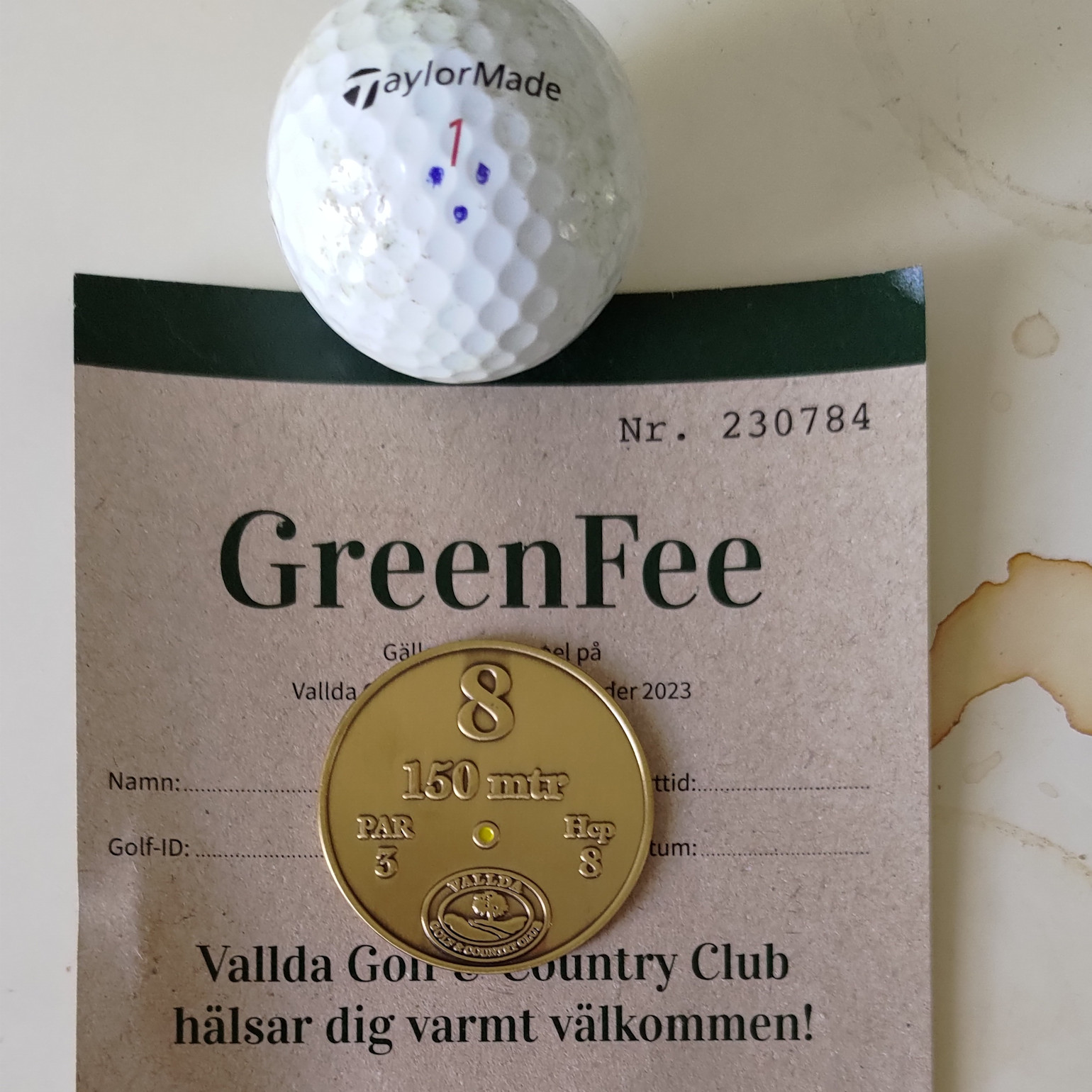 En golfbold med TaylorMade-logo, en greenfee-kvittering fra Vallda Golf & Country Club og en guldfarvet markeringsbrik til hul 8, 150 meter, par 3.