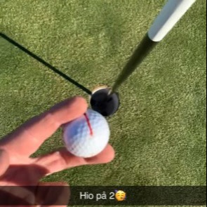 En hånd holder en golfbold tæt på et golfhul med flagstang på en grøn puttinggreen, tekst: "Hio på 2".