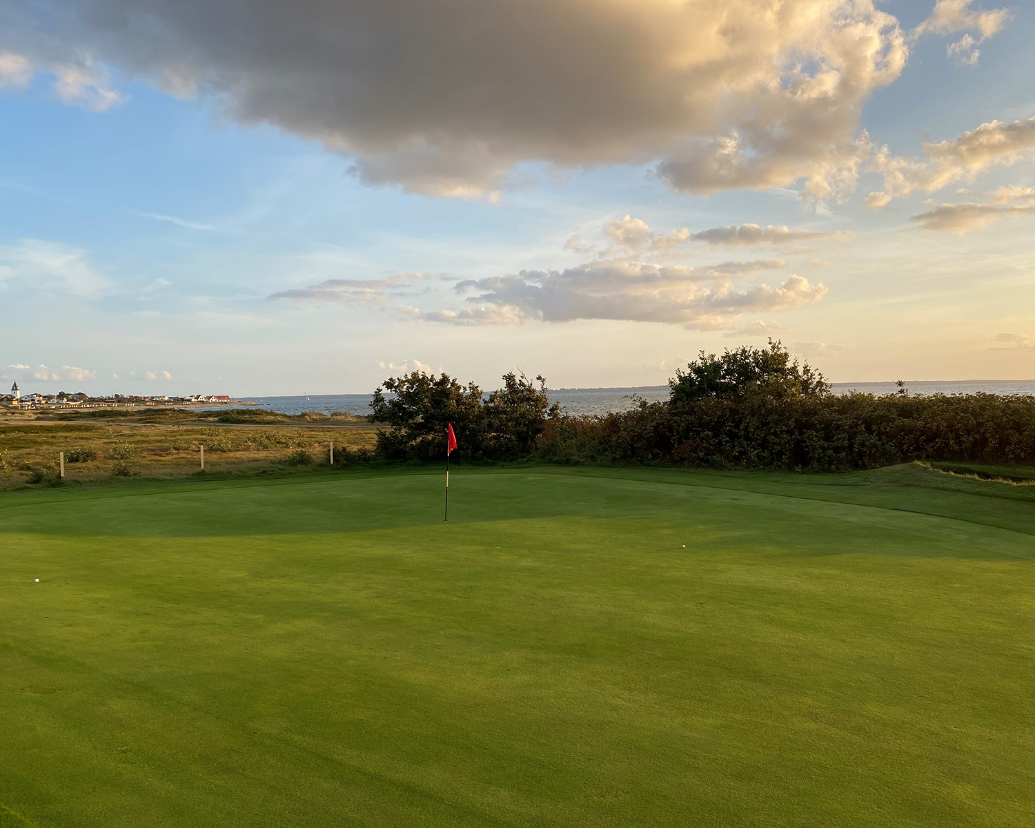 Golfgreen med flagga i mitten, omgiven av buskar och hav i bakgrunden under en delvis molnig himmel vid solnedgång.