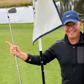 Smilende mand i mørk jakke og blå kasket holder en golfkølle og peger mod en flagstang på en golfbane.