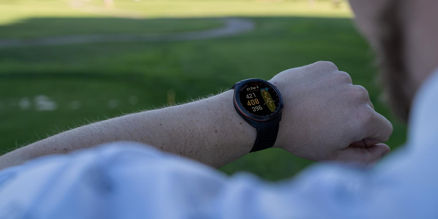 Eine Person schaut auf eine Smartwatch, die Golfdaten wie Loch, Par und Entfernung auf einem Golfplatz im Freien anzeigt.