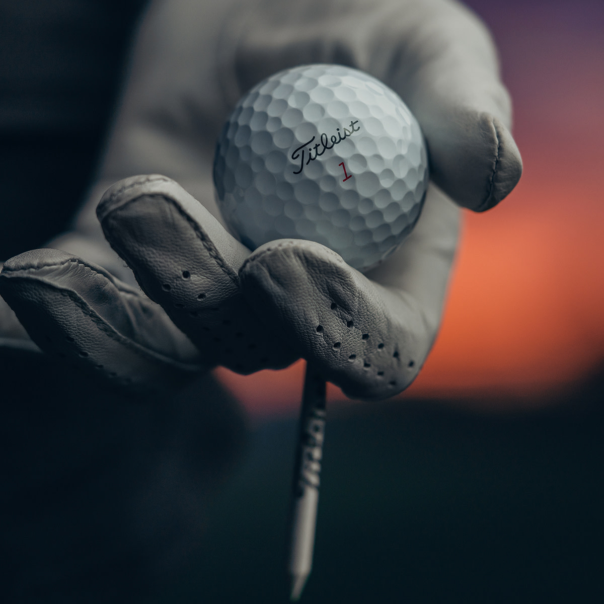 Käsi, jossa on golfhanska, pitää Titleist-golfpalloa ja tiitä epätarkkaa taustaa vasten auringonlaskussa.