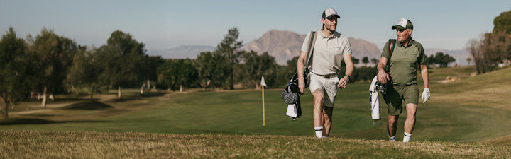 Two men are walking on a golf course by the water, both wearing caps and padded vests, one blue and one orange.