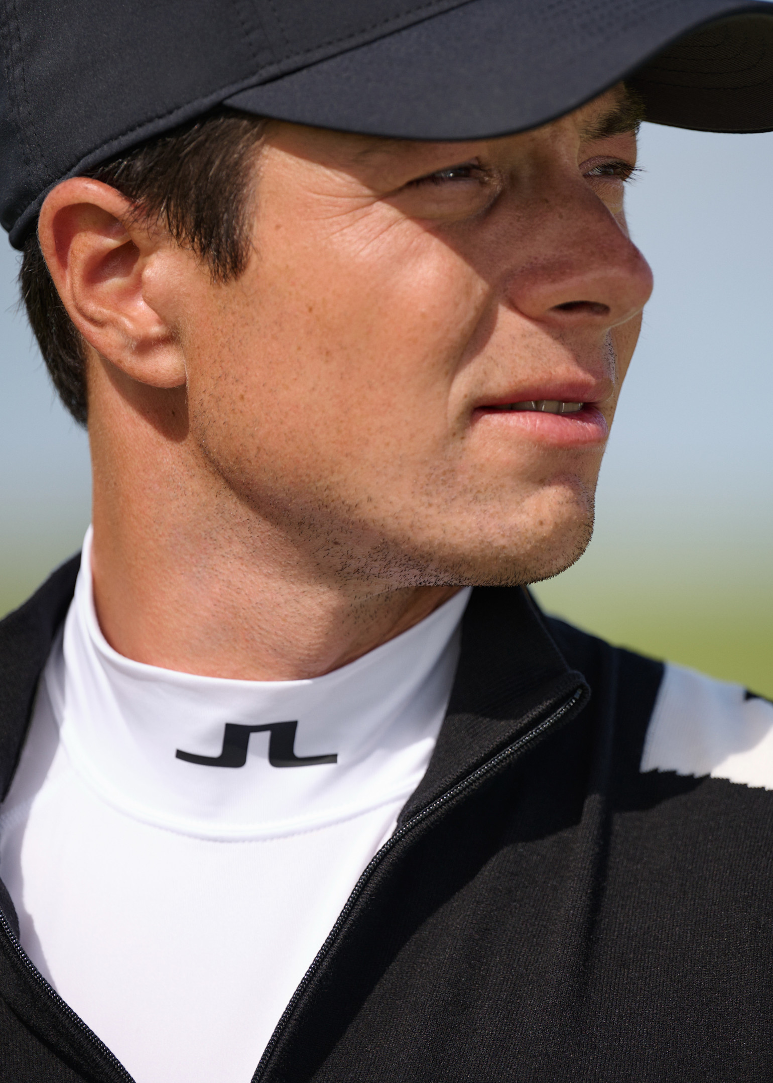 Mann mit schwarzer Kappe und schwarz-weißer Jacke blickt im Freien zur Seite, JL-Logo ist auf seinem weißen Kragen zu sehen.