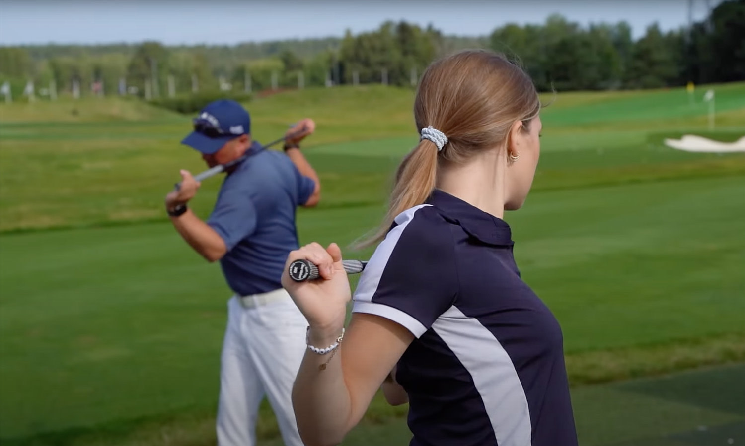 Två golfspelare värmer upp med golfklubbor över axlarna på en grön golfbana med träd i bakgrunden.