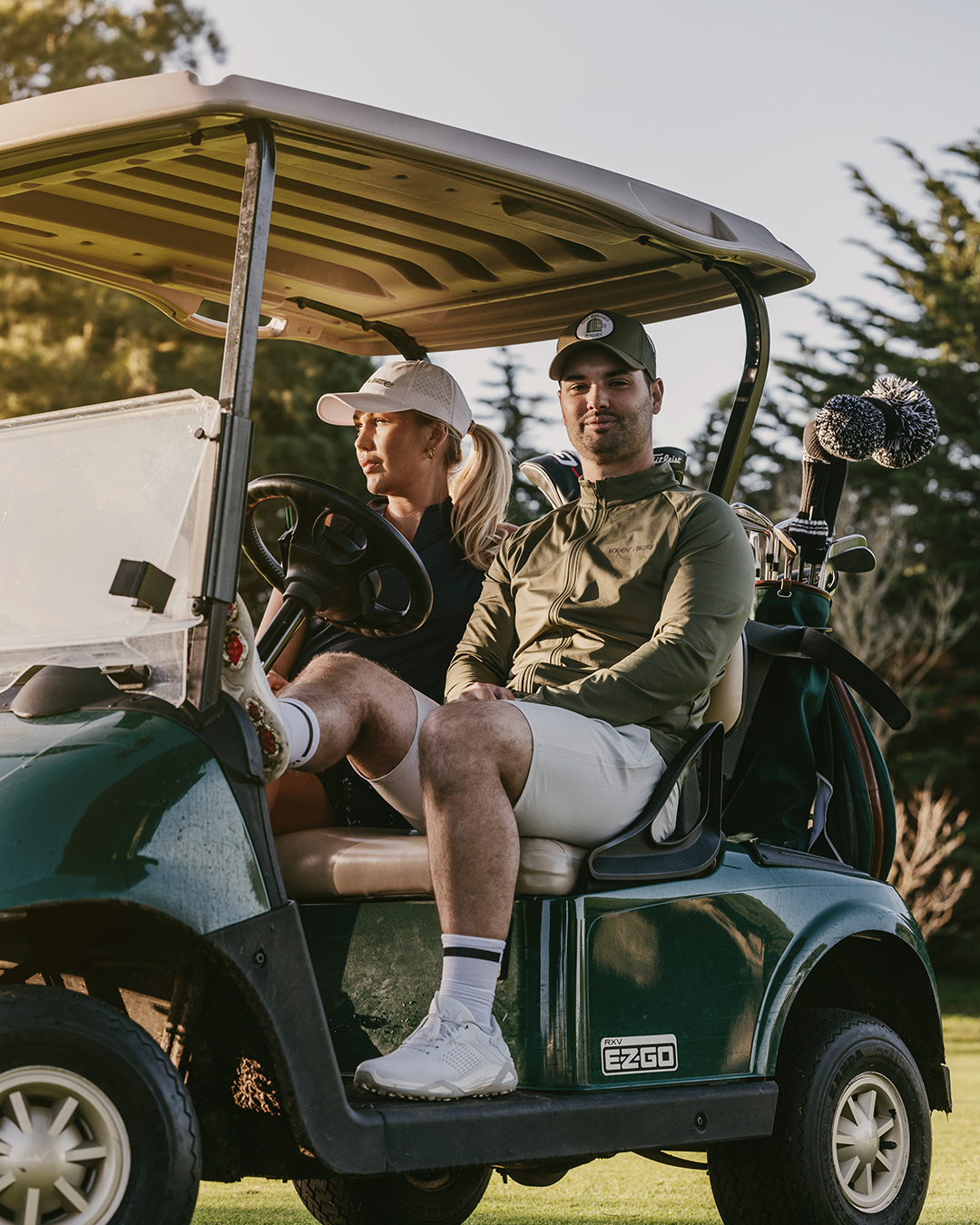 Two people are sitting in a golf buggy on a golf course, surrounded by trees, with golf clubs in the background.