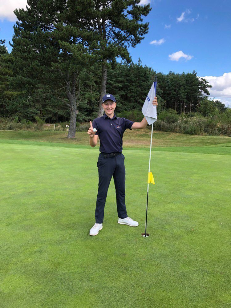 Ung man i golfkläder håller en golfboll och flagga på greenen, ler och pekar mot hålet, omgiven av träd och blå himmel.