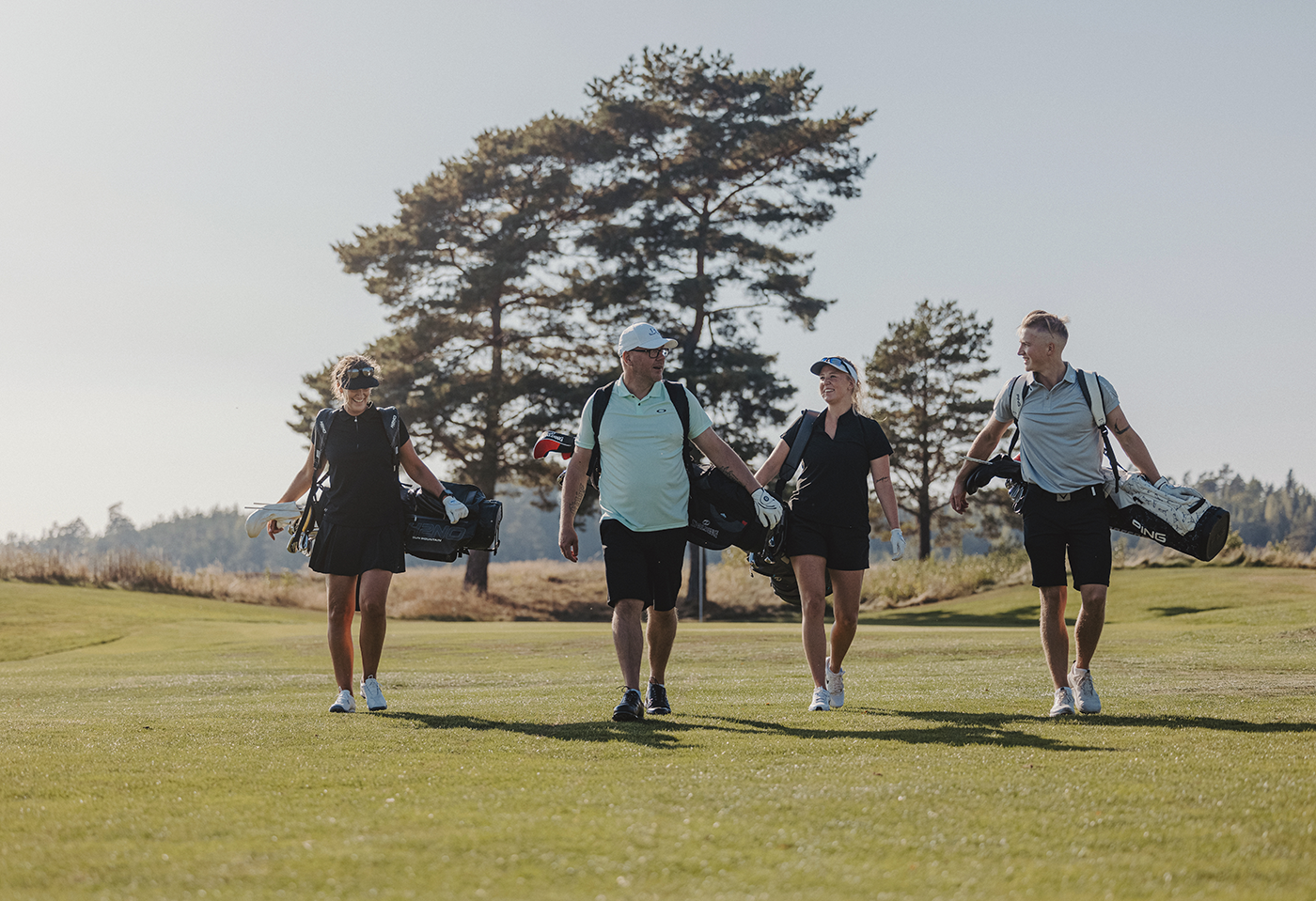 Vier Personen gehen gemeinsam auf einem Golfplatz, tragen Golfbags und lächeln einander an an einem sonnigen Tag mit Bäumen im Hintergrund.