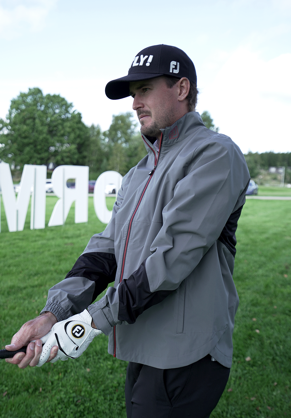 Man i grå jacka och svart keps med texten "FLY!" håller i en golfklubba på en gräsmatta med träd i bakgrunden.
