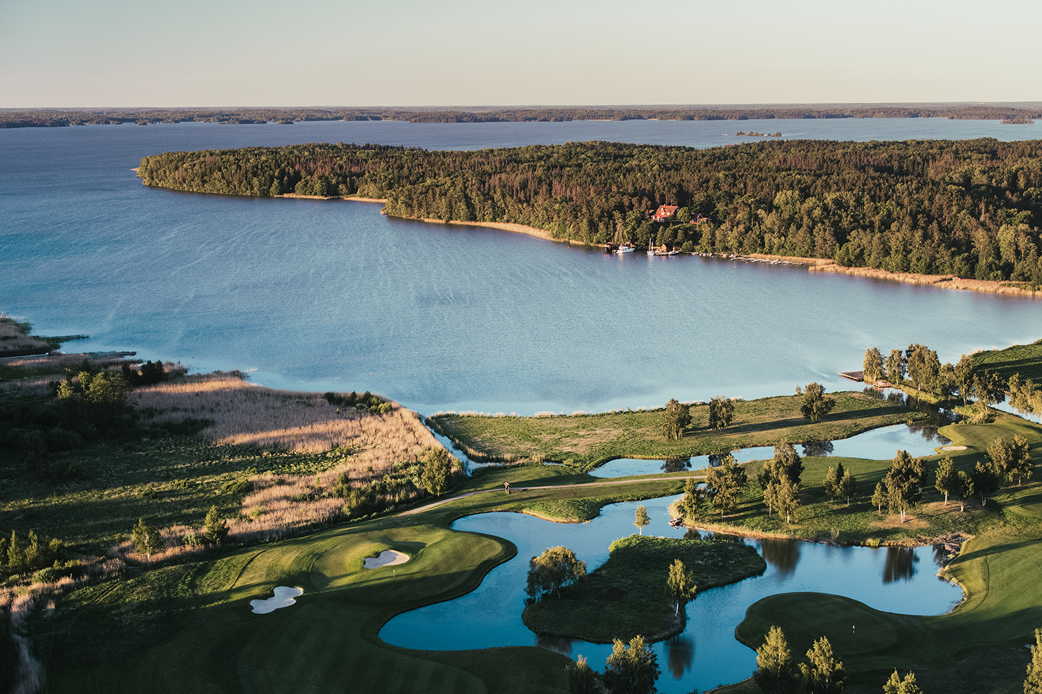 Golfbana med gröna fairways och vattenhinder i förgrunden, omgiven av sjö och skogsklädda öar i bakgrunden.