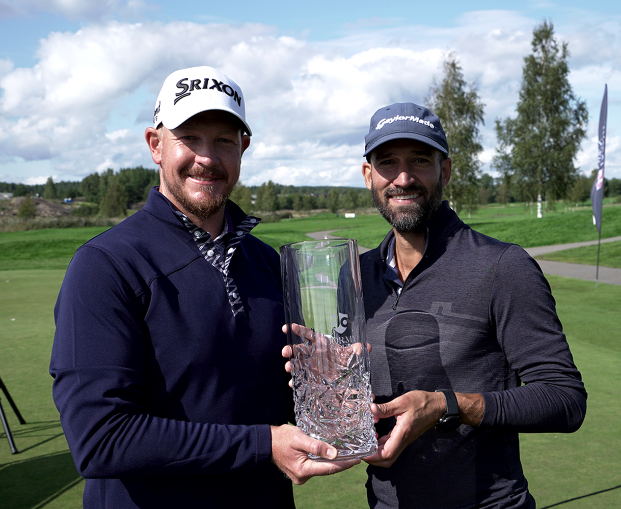 Two men in golf attire are holding a glass trophy together on a golf course on a sunny day.
