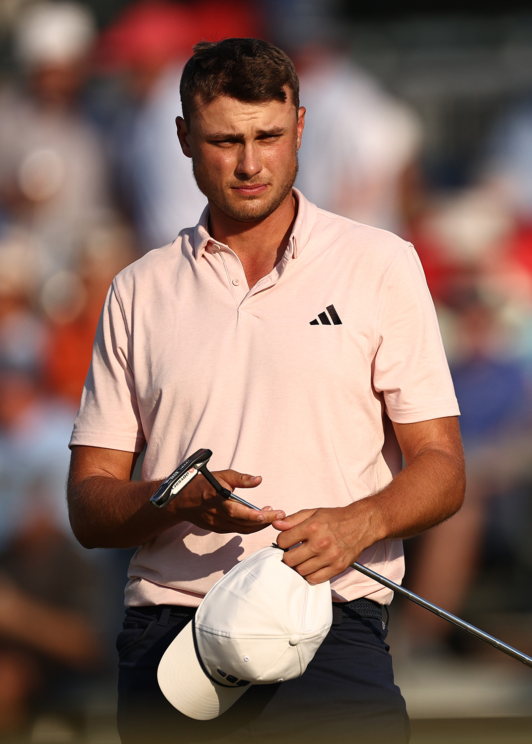 Man i ljusrosa pikétröja med Adidas-logga håller en golfklubba och vit keps, står utomhus med suddig publik i bakgrunden.