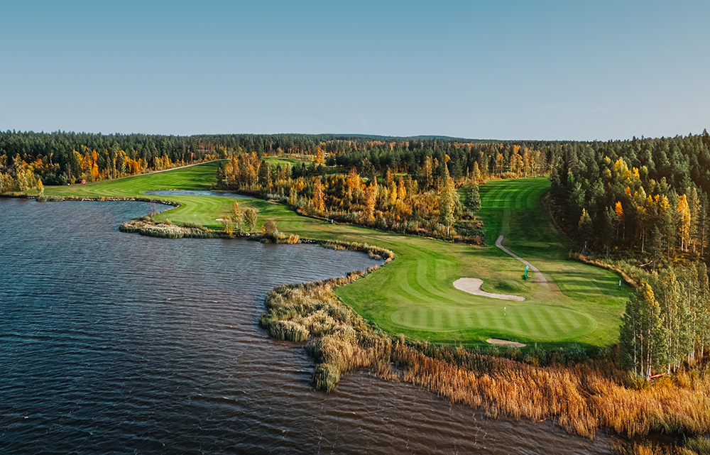 Golfbana med välklippta greener och sandbunker vid en sjö, omgiven av höstfärgade träd och skog under klar himmel.