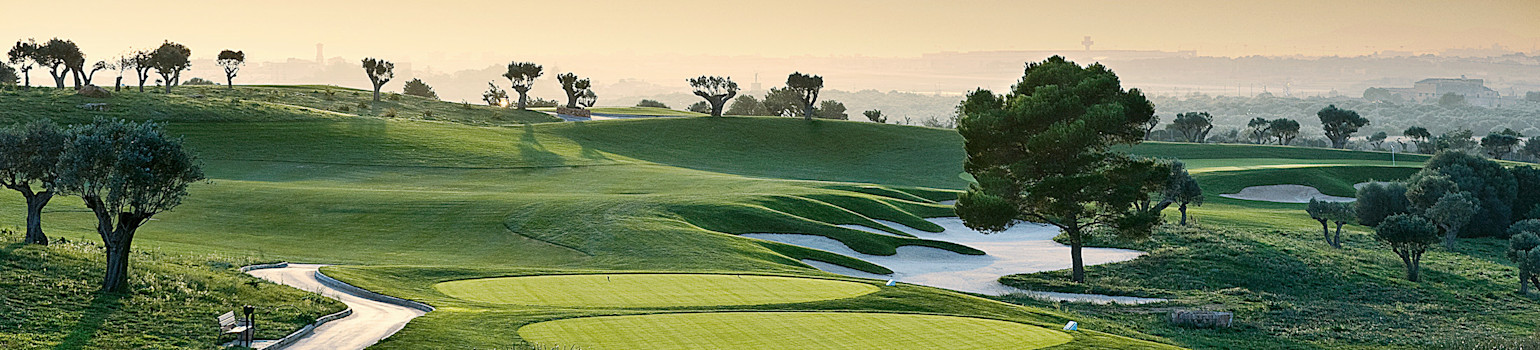 Grön golfbana med välklippta fairways och bunkrar, omgiven av träd och gräs under en dimmig morgonhimmel.